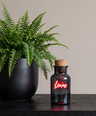 A small dark glass bottle with a cork stopper, lit with a glowing red neon-style 'love' script on the front, set on a dark red surface against a dark background with blurred cards behind it.