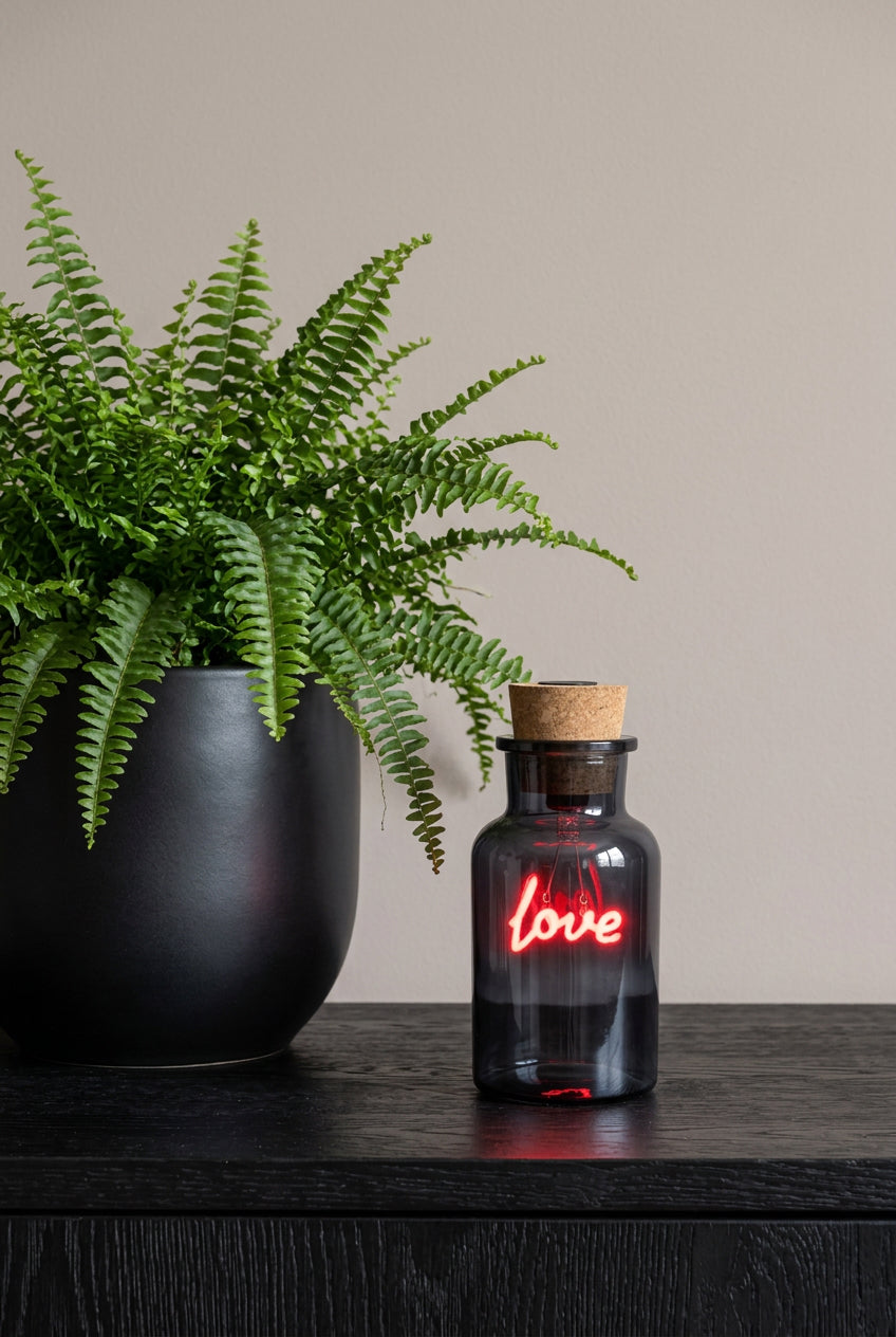 A small dark glass bottle with a cork stopper, lit with a glowing red neon-style 'love' script on the front, set on a dark red surface against a dark background with blurred cards behind it.