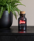 A small dark glass bottle with a cork stopper and a glowing red neon-style 'love' sign, placed on a dark wooden surface beside a large black planter with green ferns.