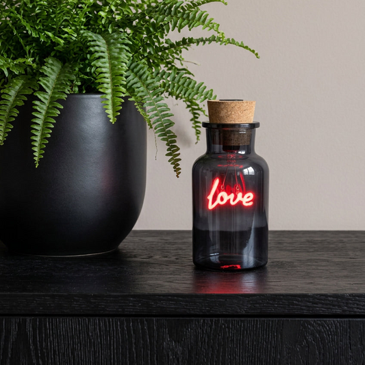 A small dark glass bottle with a cork stopper and a glowing red neon-style 'love' sign, placed on a dark wooden surface beside a large black planter with green ferns.