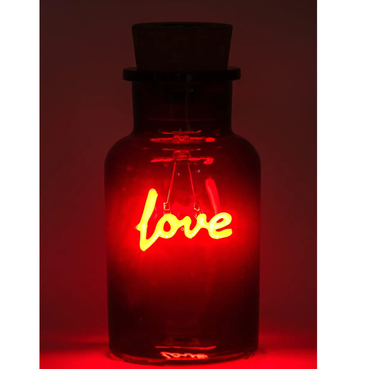 A dark glass bottle with a cork stopper, illuminated from within by a glowing red neon-style 'love' sign, set against a dark red background.