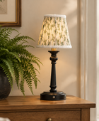 A classic table lamp with a black turned-wood style base and a beige floral-patterned fabric shade with a white lace trim, sitting on a wooden table next to a potted fern.