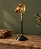 A vintage-style brass and black table lamp with a small lit candle holder and oval mirror, displayed on a rustic wooden table next to an old book and a potted olive branch against a dark green wall.