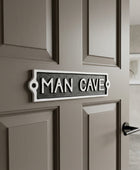 A rectangular metal door sign reading 'MAN CAVE' in raised white letters on a dark textured background, mounted on a taupe paneled door.