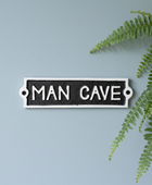A rectangular black and white metal sign with the words 'MAN CAVE' in white capital letters, mounted against a light blue wall with green fern leaves on the right side.