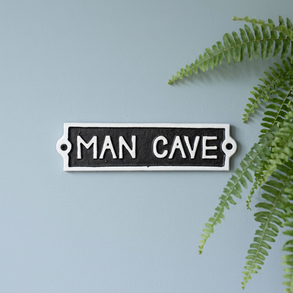 A rectangular black and white metal sign with the words 'MAN CAVE' in white capital letters, mounted against a light blue wall with green fern leaves on the right side.