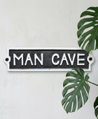 A rectangular cast iron sign with the words 'MAN CAVE' in raised white lettering on a dark textured background, mounted against a rustic wooden surface.