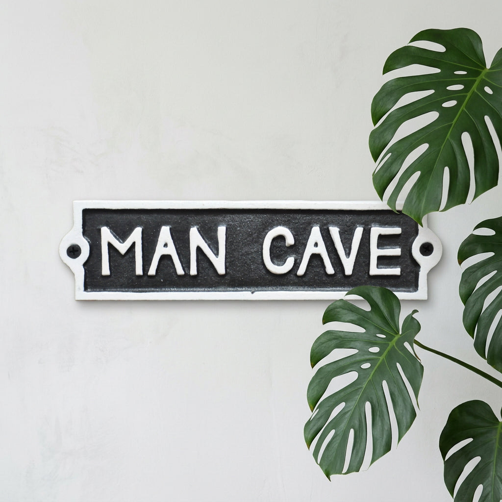 A rectangular cast iron sign with the words 'MAN CAVE' in raised white lettering on a dark textured background, mounted against a rustic wooden surface.