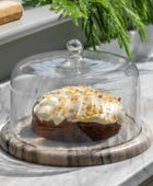 Decorative cake with white frosting and gold sprinkles under a glass cloche on a marble surface.