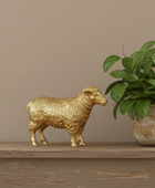 A small metallic gold sheep figurine standing on a rustic wooden shelf next to a green potted plant against a warm beige wall.