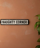 Sign with 'NAUGHTY CORNER' text on a textured wall with a plant.
