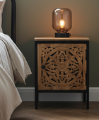 Wooden nightstand with decorative carvings and a lit table lamp on a bed side.