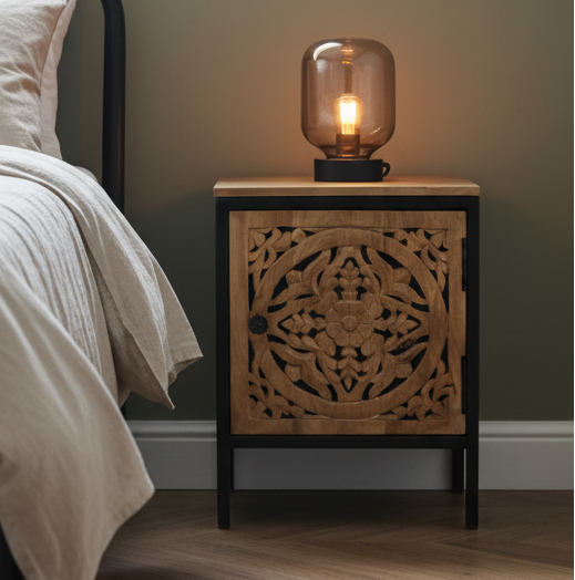 Wooden nightstand with decorative carvings and a lit table lamp on a bed side.