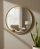 Round wall mirror with a thin gold frame mounted on a beige wall, reflecting a softly lit living room interior. A potted snake plant is visible in the lower right corner.