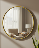 Round wall mirror with a thin gold frame, mounted on a beige wall. The mirror reflects a softly lit living room with a chair, side table, and lamp, while a green snake plant is visible in the foreground.