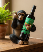 A novelty figurine of a seated chimpanzee holding a large wine bottle with a green label reading 'Malbec' and 'Intense & Rich.' The figurine is placed on a wooden surface next to a potted fern in a real indoor setting.