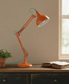Orange desk lamp on a wooden surface with a plant and book in a room with a window.