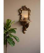 An ornate gold wall mirror with a decorative corset-style frame and matching shelf, mounted on a pale pink paneled wall with a blurred green plant in the foreground.