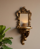An ornate gold wall sconce with a decorative mirror and shelf, mounted on a beige wall with a green plant in the foreground.