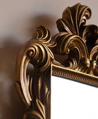 Close-up of an ornate gold mirror frame with carved baroque-style scrollwork and a reflective white mirror surface, mounted against a beige wall.