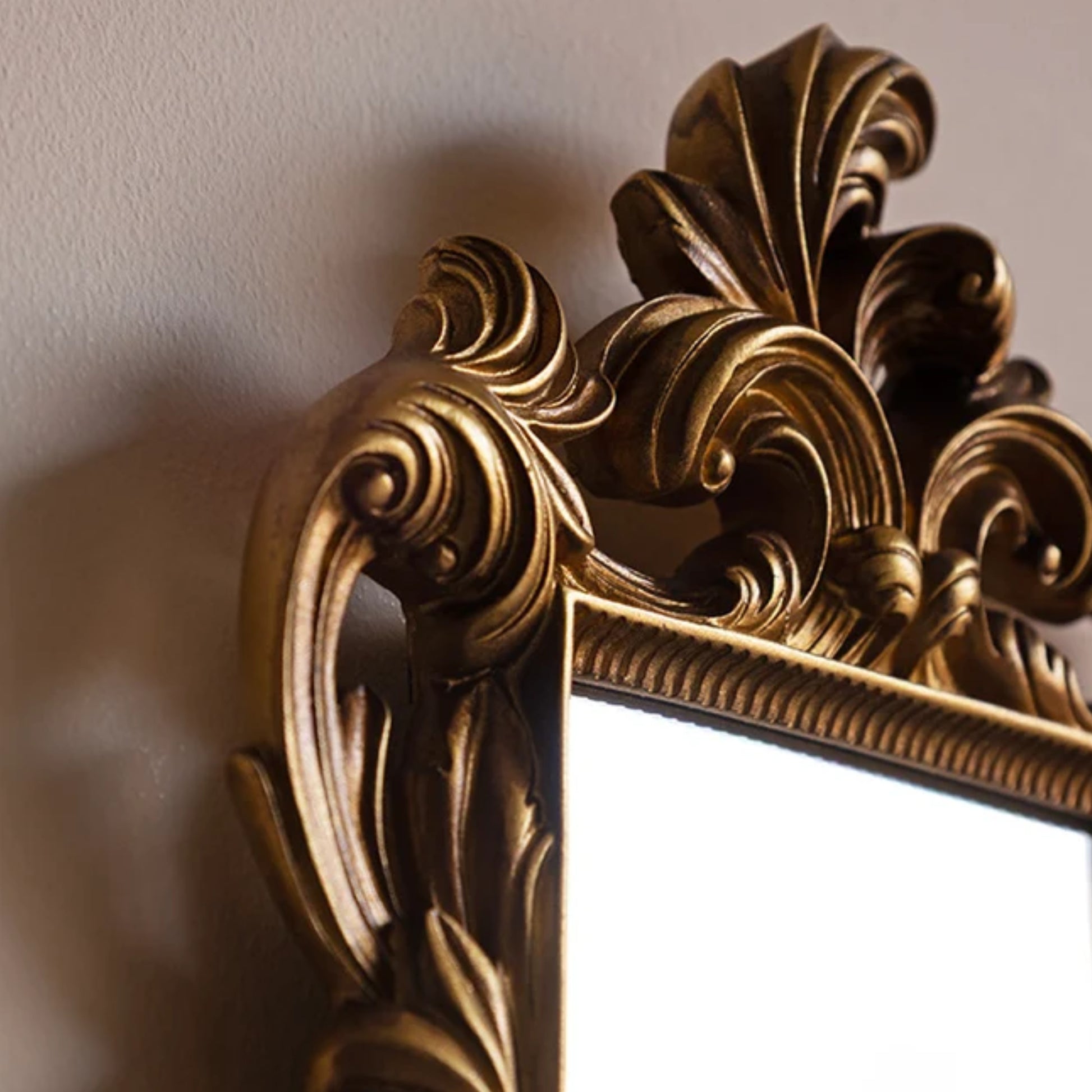 Close-up of an ornate gold mirror frame with carved baroque-style scrollwork and a reflective white mirror surface, mounted against a beige wall.