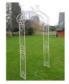 Decorative white metal garden archway on a grassy lawn with trees and a building in the background.