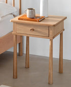 Wooden side table with a drawer next to a bed, featuring a mug and book on top.