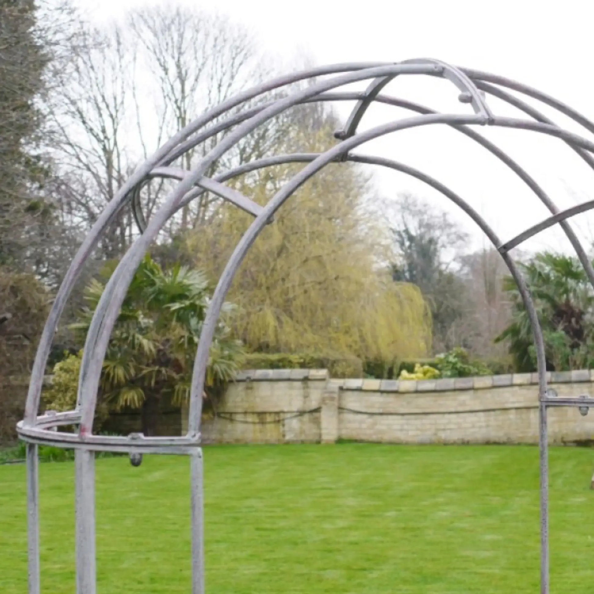 Metal garden archway in a garden setting with trees and a stone wall in the background.