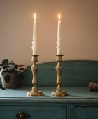 Two lit candles in ornate brass candlesticks on a wooden surface with a plant and book in the background.