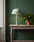 A light sage green table lamp with a sculptural, tiered spiral base sits on a rustic wooden console table next to a potted fern against a dark green wall.