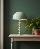 A mint green table lamp with a ribbed cylindrical base sits on a wooden console table next to a potted fern against a dark green wall.