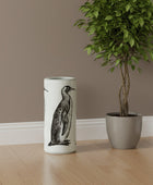 Cylindrical vase with penguin design next to a potted plant on a wooden floor.