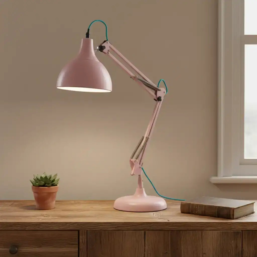 Pink desk lamp on a wooden surface with a plant and book in a room with a window.