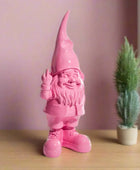 Pink gnome figurine waving next to a small potted plant on a wooden surface with a light purple wall background.