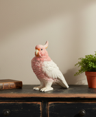 Decorative parrot figurine on a wooden surface with a plant and book in the background.