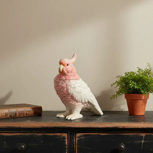 Decorative parrot sculpture on a wooden surface with a plant and book in the background.