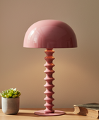 Pink table lamp with a textured base on a wooden surface next to a small plant and book against a beige wall.
