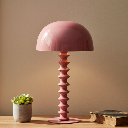 Pink table lamp with a textured base on a wooden surface next to a small plant and book against a beige wall.