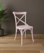 Pink chair with cross-back design on a wooden floor next to a plant against a gray wall.