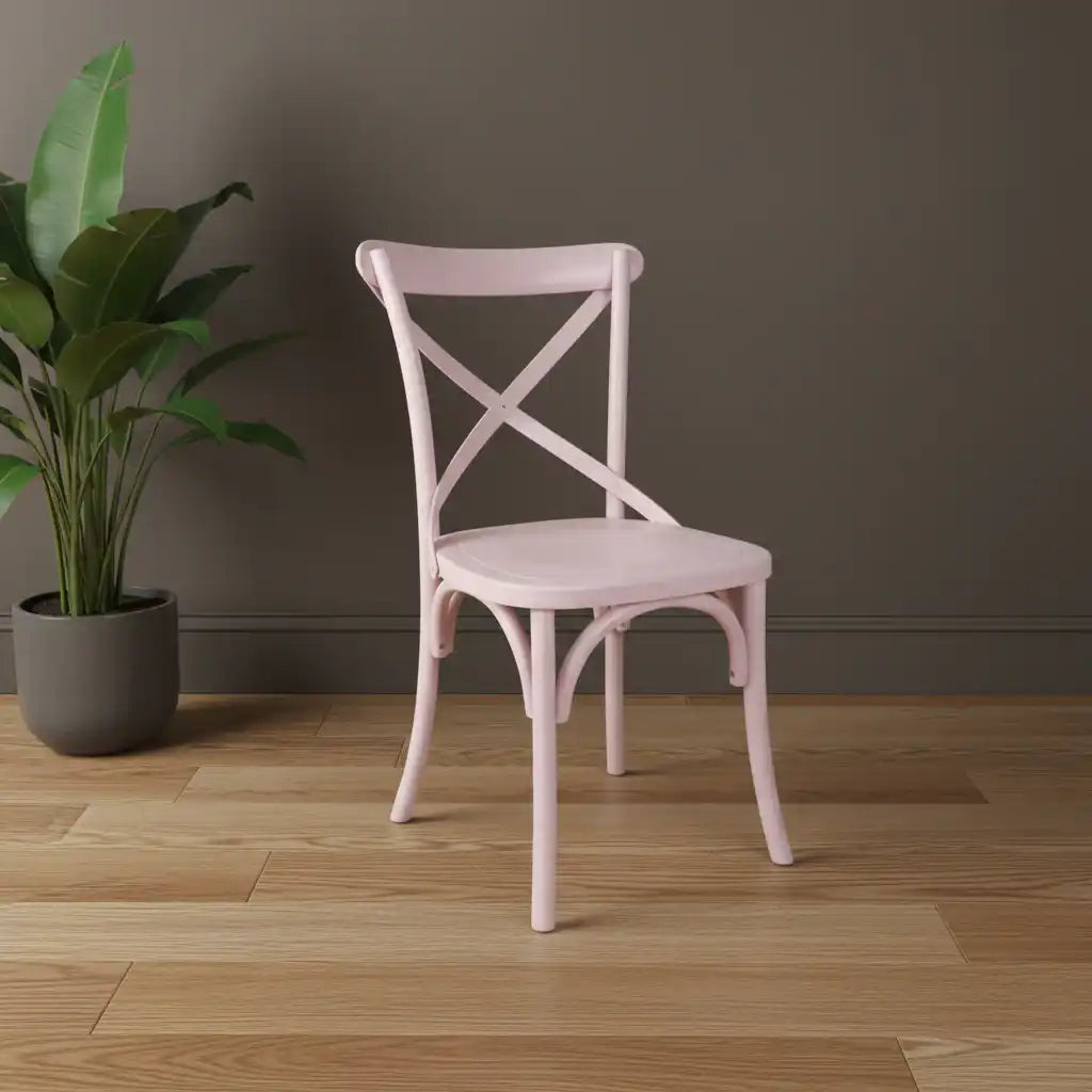 Pink chair with cross-back design on a wooden floor next to a plant against a gray wall.