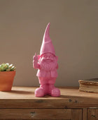 Pink gnome figurine on a wooden surface with a potted plant and book in the background.