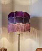 A floor lamp with a purple velvet shade featuring a scalloped edge and pink fringe, mounted on a gold-toned metal pole. It is styled in a cozy living room setting next to a window and a sofa.