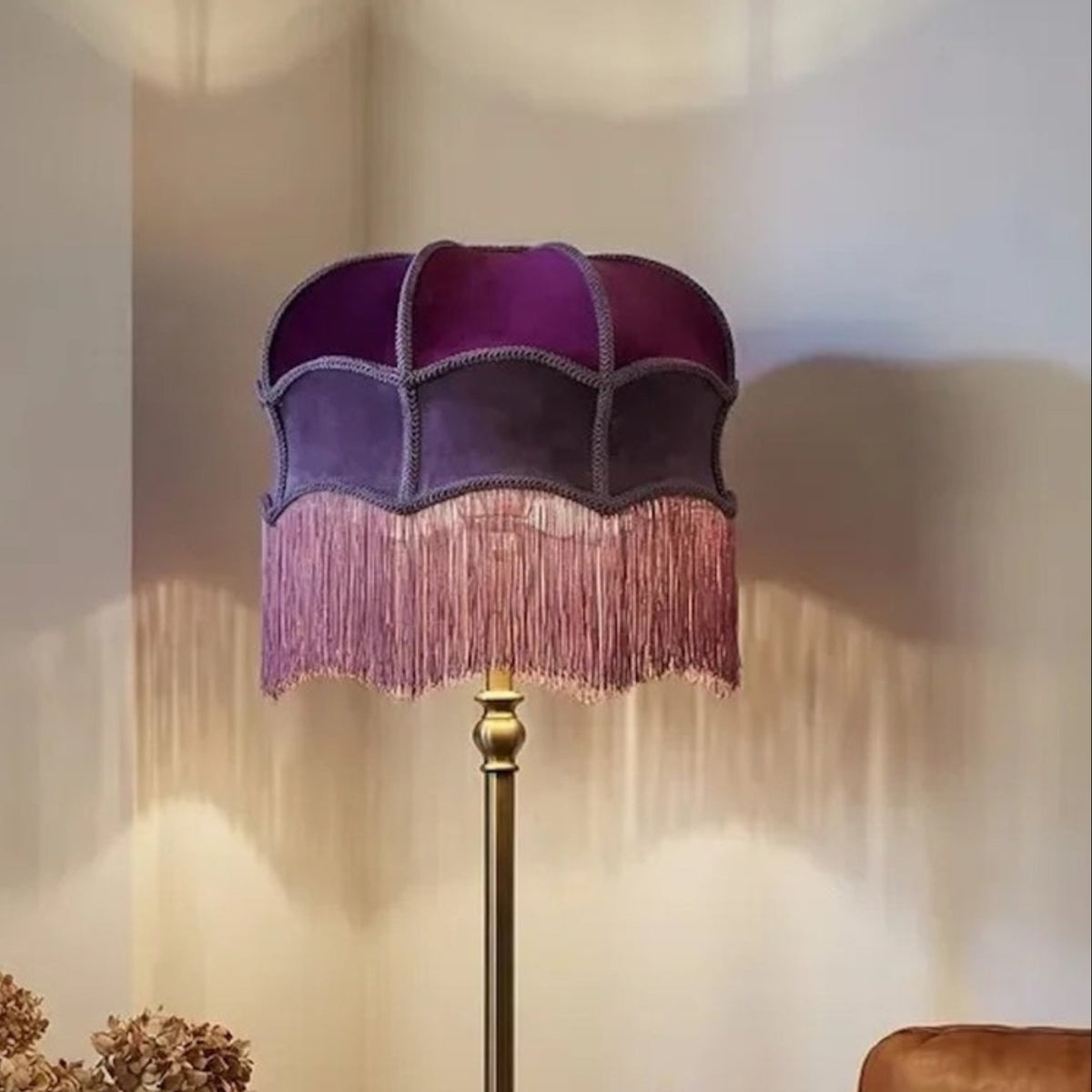 A floor lamp with a purple velvet shade featuring a scalloped edge and pink fringe, mounted on a gold-toned metal pole. It is styled in a cozy living room setting next to a window and a sofa.