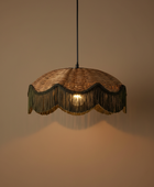 A hanging pendant light with a woven natural-fiber shade and dark fringe, glowing with a warm light against a plain beige wall.