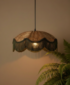 A woven pendant light with a scalloped fringe shade, hanging against a warm beige wall with a green fern in the foreground.