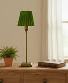 Green lamp on a wooden table with a plant and book, in a room with a window and beige wall.