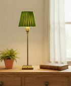 Green lamp on a wooden table with a plant and book in a room with a window.