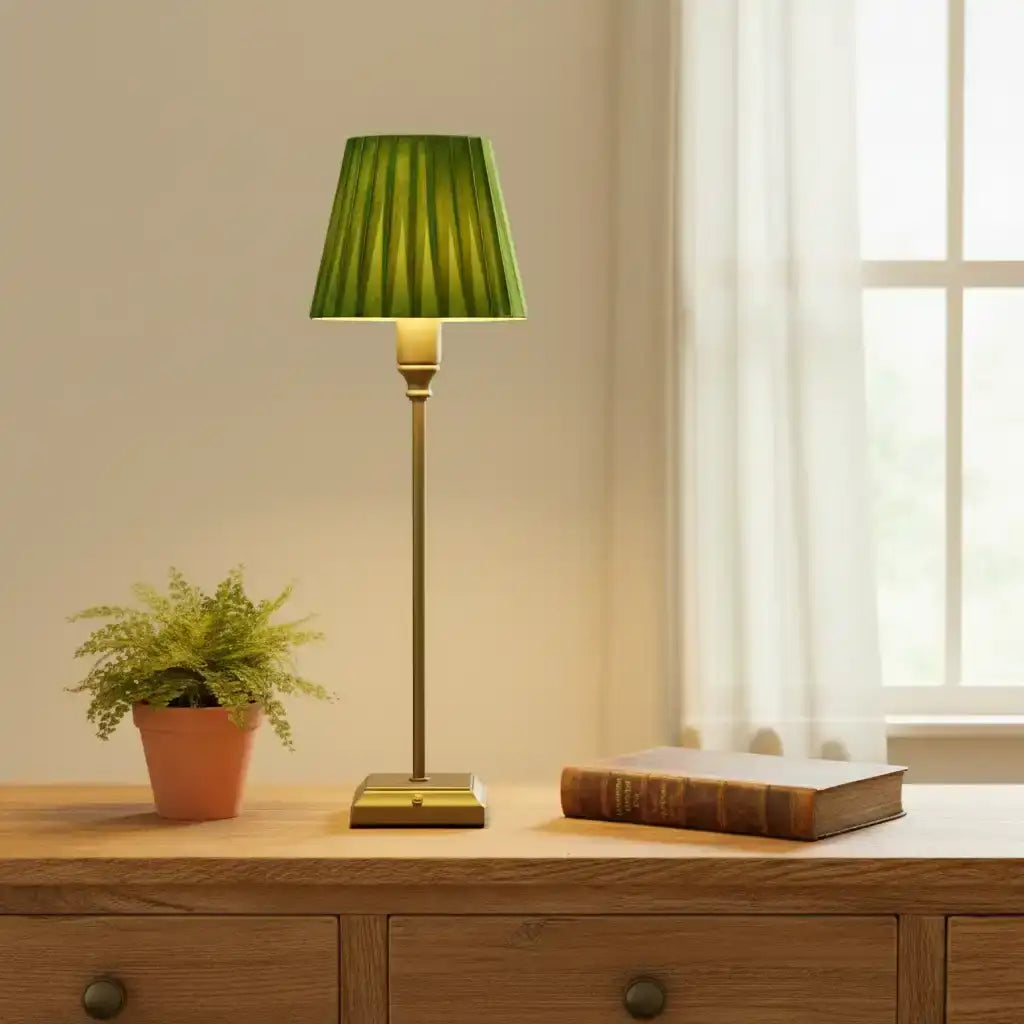 Green lamp on a wooden table with a plant and book in a room with a window.