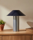 Table lamp with black shade and striped base on a wooden surface next to a plant and book.