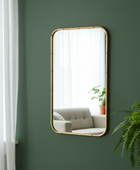Rectangular mirror with gold frame on a green wall, reflecting a living room with a sofa and plant.